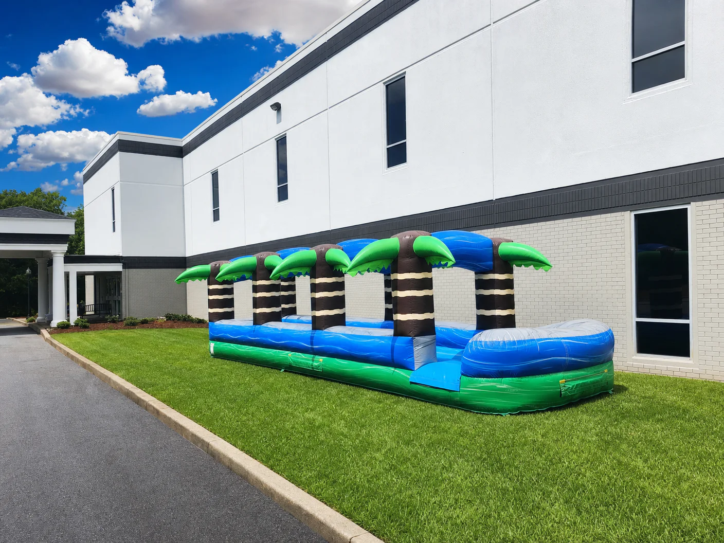 Wow Inflatables long tropical palm tree inflatable slip-n-slide with four striped palm tree pillars and blue green wave patterns set up on green grass beside building in Perry Georgia — slip and slide rental Middle Georgia