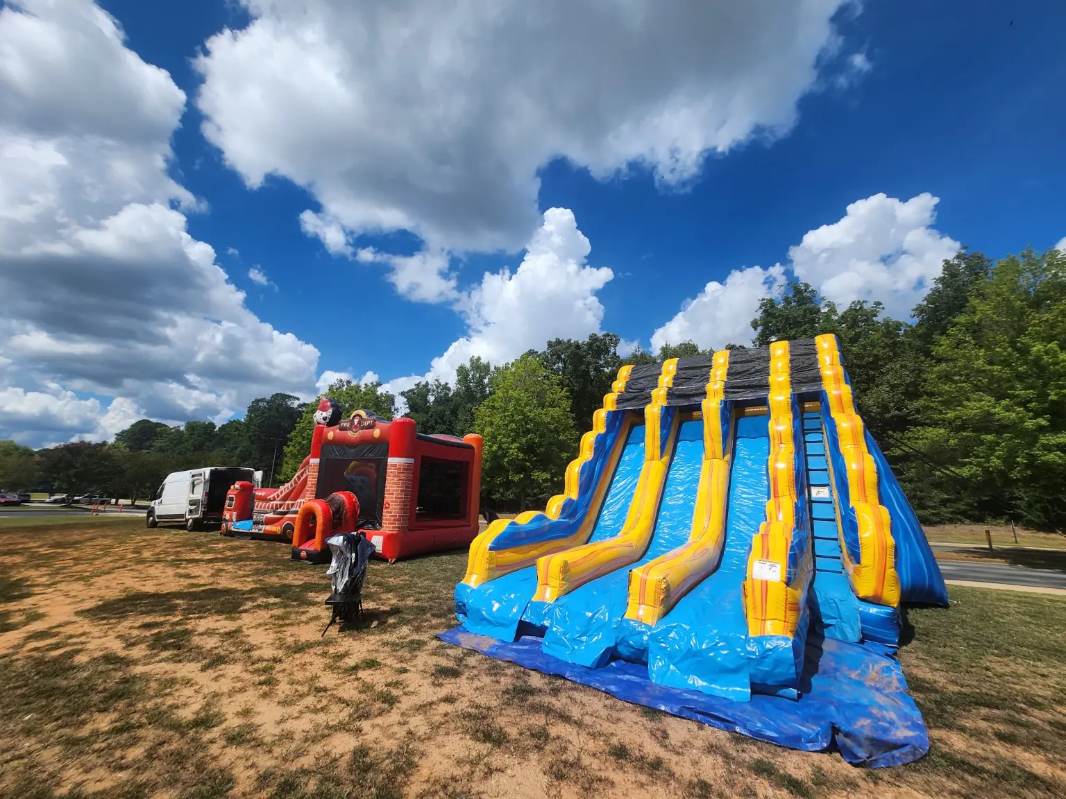 Yellow and blue triple-lane inflatable slide beside a red Fire House bounce and slide combo on grass